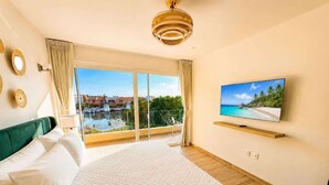 A bedroom with a large bed, a TV on the wall, and a balcony with a view of the sea. - Canal View | 2 King Beds | 7 min to Beach (Puerto Aventuras)