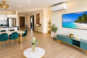 A modern living room with a dining area, a large flat-screen TV, and a view of a beach. - Luxury 3BR Canal View Condo Near Beach (Puerto Aventuras)