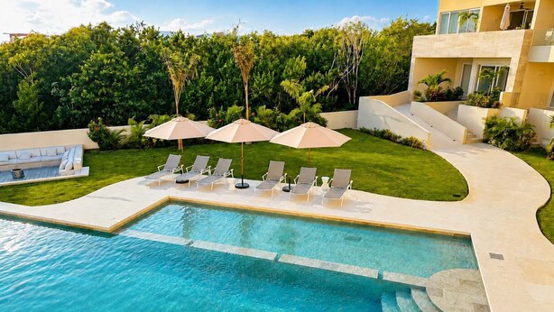 A swimming pool with lounge chairs and umbrellas, surrounded by a well-maintained lawn and a building with a modern design. - Canal View | 2 King Beds | 7 min to Beach (Puerto Aventuras)