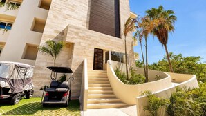 A modern building with a staircase leading to the entrance, surrounded by palm trees and a covered vehicle. - Canal View | 2 King Beds | 7 min to Beach (Puerto Aventuras)