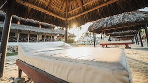 Una cama frente al mar, bajo un techo de paja, con una playa arenosa y palmeras al fondo.