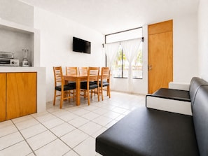 Un espacio habitable compacto con kitchenette, una zona de comedor con sillas de madera, un sofá negro y un televisor montado en la pared.