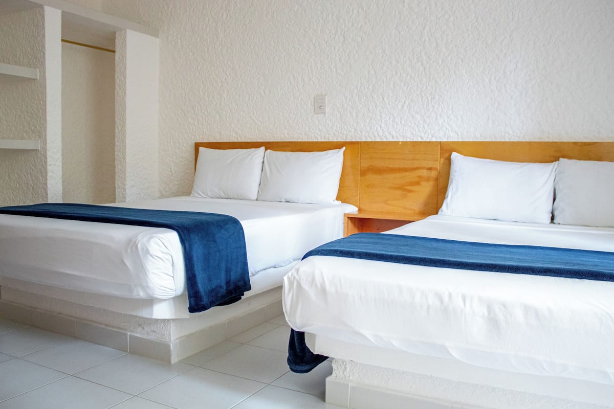 Dos camas individuales con ropa blanca y mantas azules, un cabecero de madera y una pared blanca con textura.