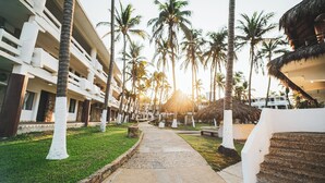 Un resort con un sendero bordeado de palmeras, una construcción de techo de paja y un edificio de varios pisos con balcones.