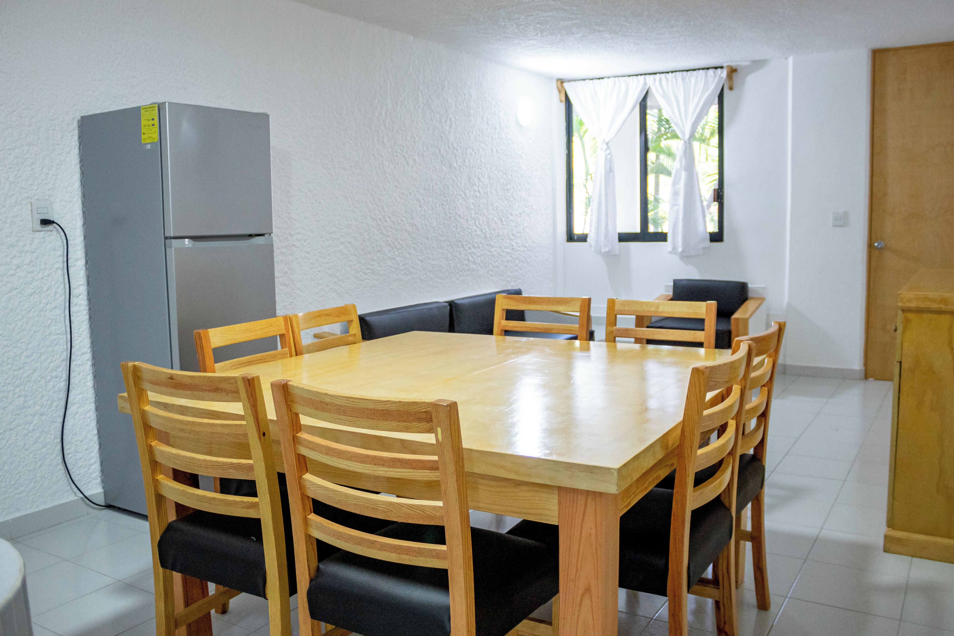 A dining area with a rectangular wooden table surrounded by six chairs, a grey refrigerator, and a window with white curtains.