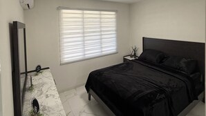 Un dormitorio con cama, mesita de noche, espejo, ventana con persianas y luz en el techo.