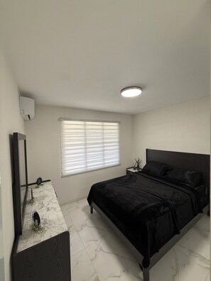 A bedroom with a bed, a nightstand, a mirror, a window with blinds, and a ceiling light. - 3-bedroom apartment in adorable Isabela with cool AC (Isabela)