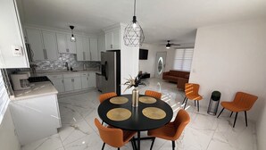 Una cocina moderna con armarios blancos, un comedor con una mesa negra y sillas naranjas, y una sala de estar con un sofá marrón.