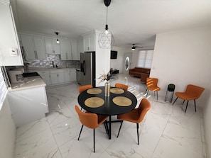 Una cocina moderna con armarios blancos, un comedor con una mesa negra y sillas naranjas, y una sala de estar con un sofá marrón.