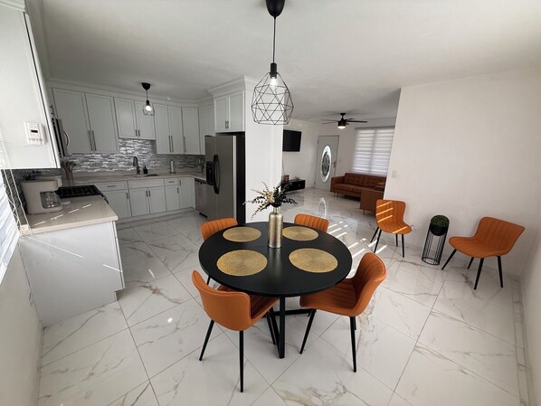 A modern kitchen with white cabinets, a dining area with a black table and orange chairs, and a living room with a brown sofa.