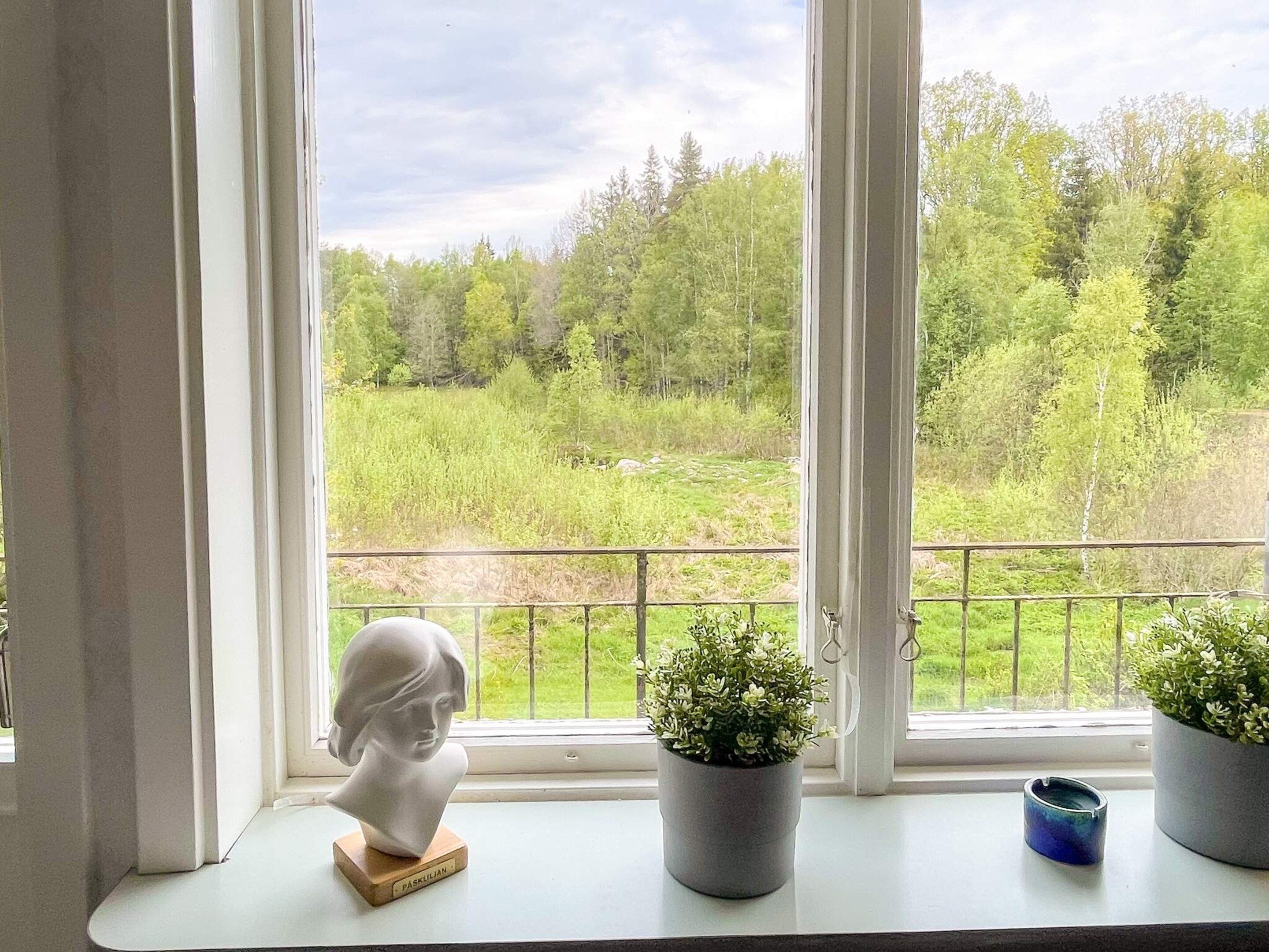 A window with a bust sculpture, two potted plants, and a view of a green landscape.