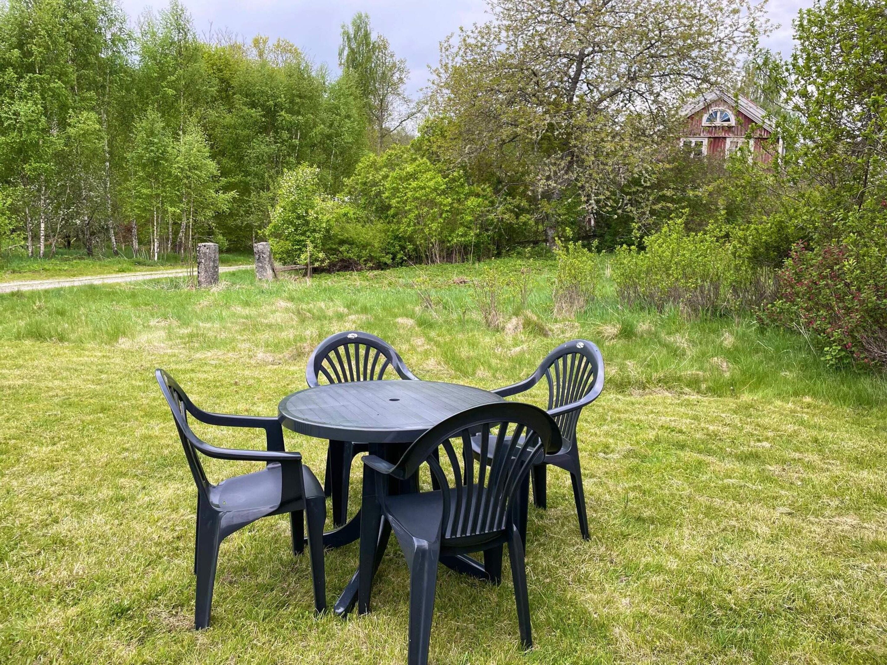 A round table with four chairs in a grassy outdoor area, surrounded by trees and a building in the background.