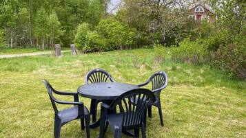 A round table with four chairs in a grassy outdoor area, surrounded by trees and a building in the background.
