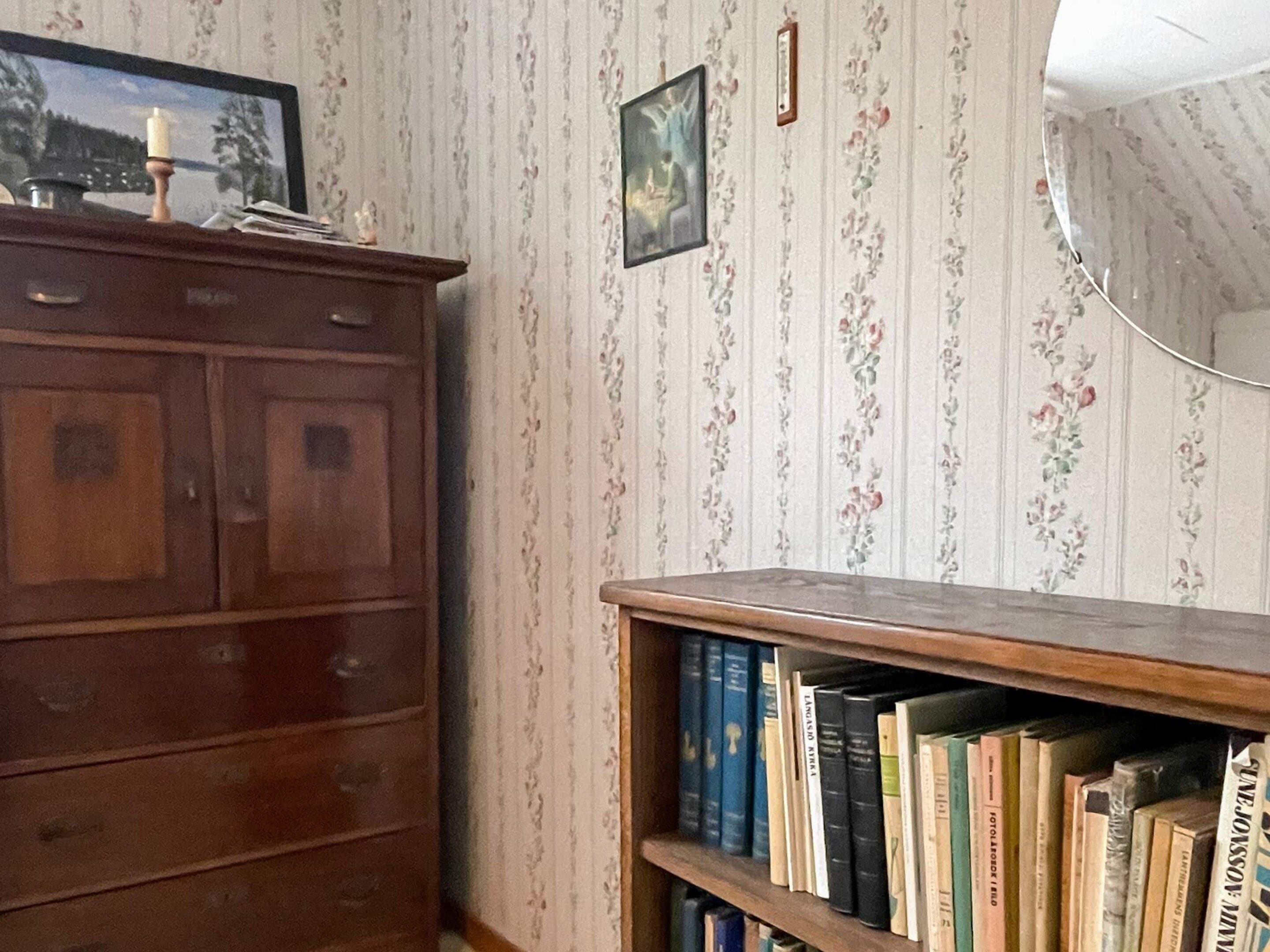 A room with a wooden cabinet, a bookshelf, and a wall with floral wallpaper.