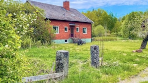 Ein rotes Backsteinhaus mit geneigtem Dach und Schornstein, umgeben von einer Wiese und Bäumen.