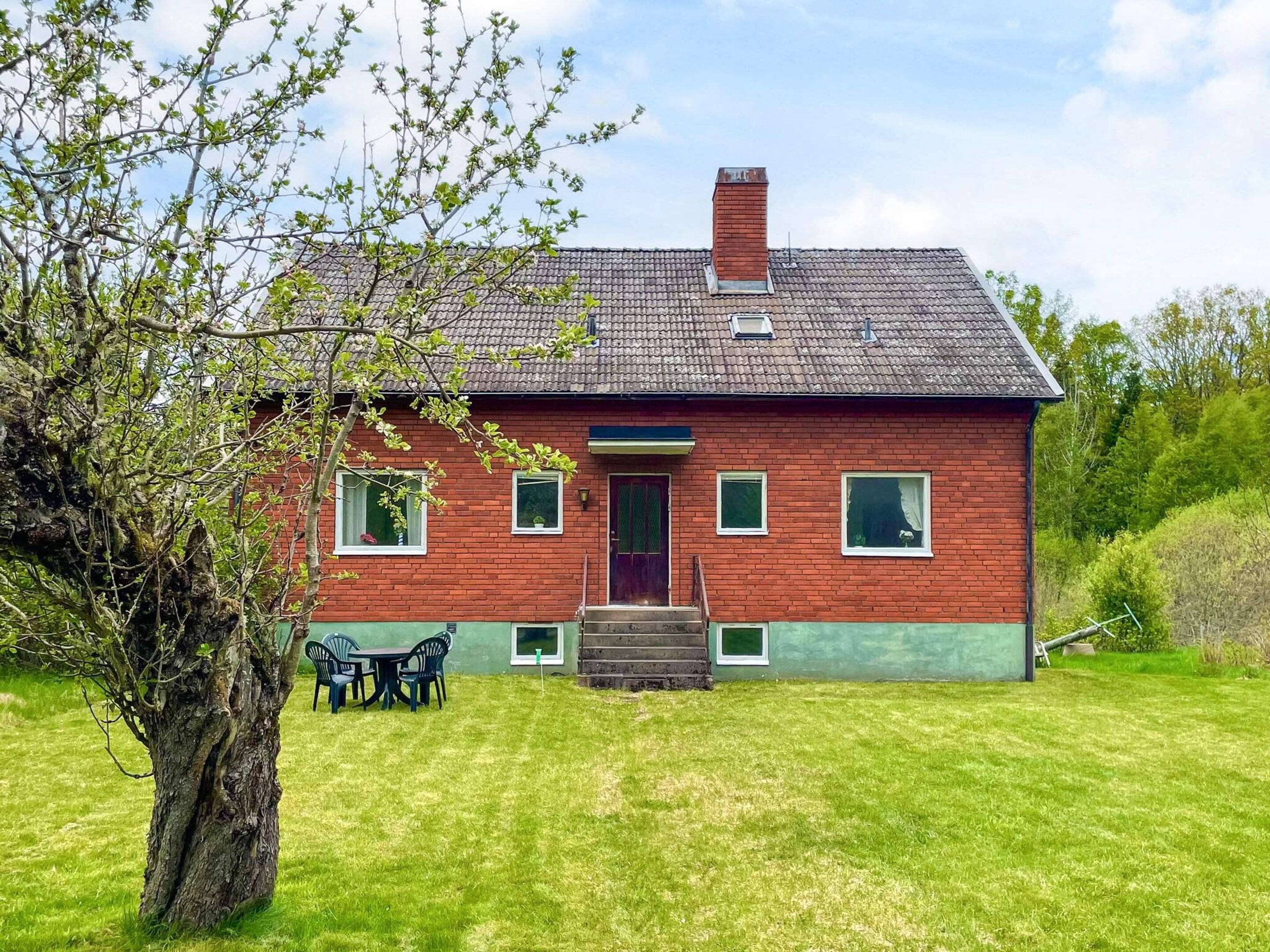 A red brick house with a green lawn, a wooden door, and a small porch with chairs.