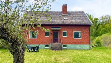 A red brick house with a green lawn, a wooden door, and a small porch with chairs.