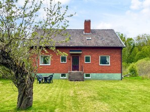 A red brick house with a green lawn, a wooden door, and a small porch with chairs. - 5 Person Holiday Home in Emmaboda (Emmaboda)