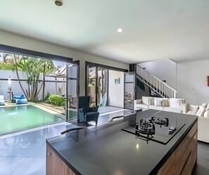 A modern kitchen with a central island, a dining area with a sofa, and a staircase leading to an upper level. - Cozzy Villa Lingkar 89 A Canggu Bali (Canggu)