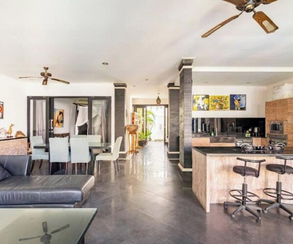 Een moderne woonkamer met een bank, een eethoek, een keuken met barstoelen en een plafondventilator.