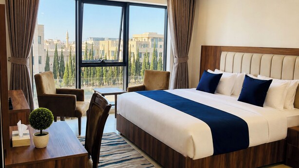Une chambre d’hôtel avec un lit, un bureau, une chaise, une petite plante en pot et une vue sur les immeubles à travers de grandes fenêtres.