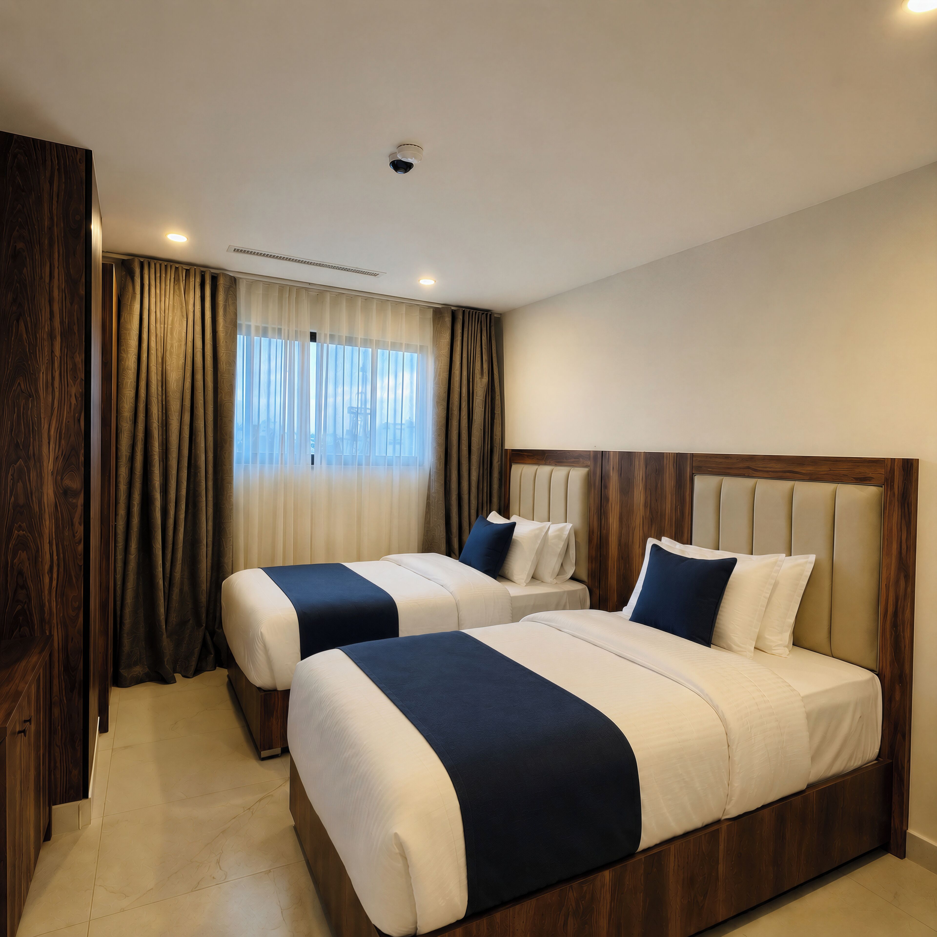 A hotel room with two beds, a wooden headboard, a large window with curtains, and a ceiling light.