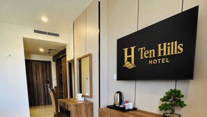 Un comptoir d’accueil d’hôtel avec une enseigne indiquant « Ten Hills Hotel », un comptoir en bois, une plante en pot, une machine à café et une télécommande.
