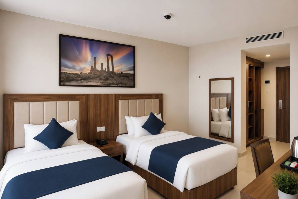 Habitación de hotel con dos camas, una mesita de noche, un escritorio, un espejo y un cuadro con ruinas antiguas.