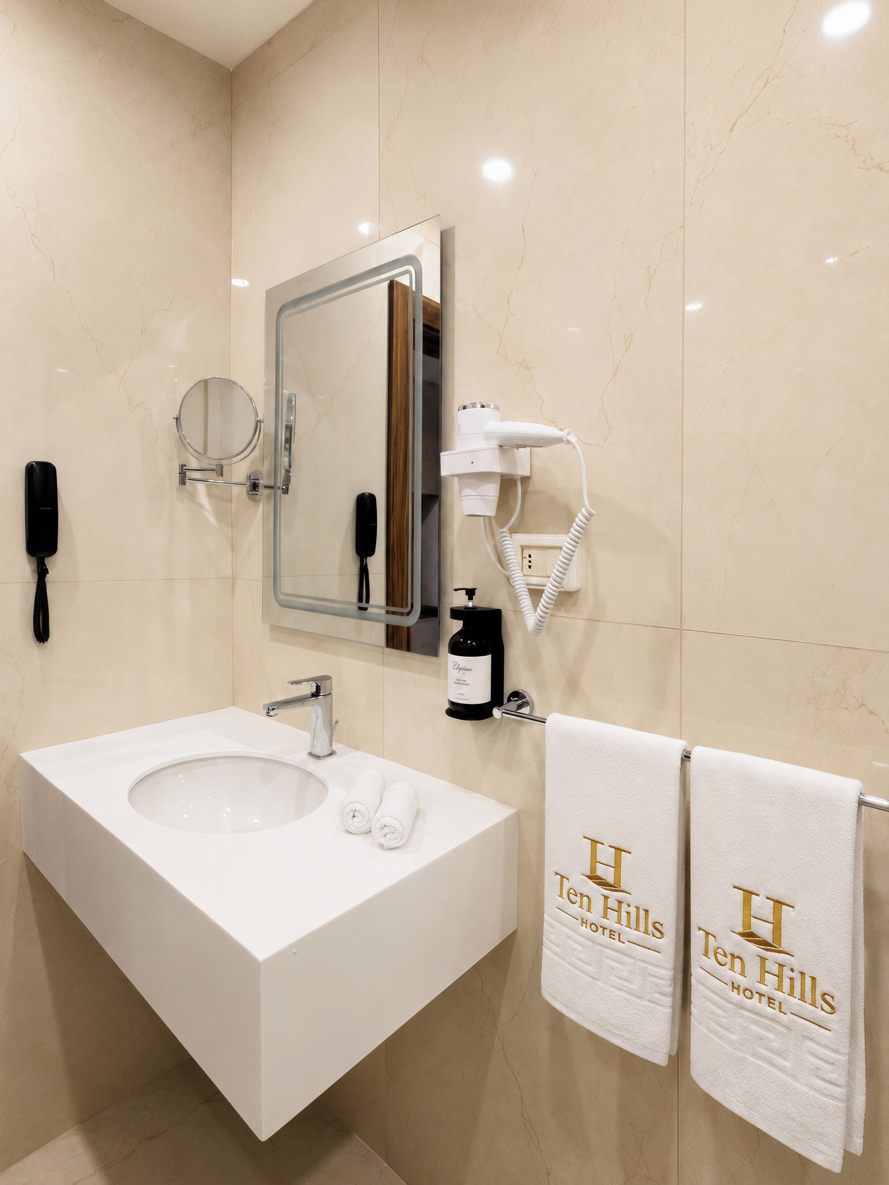 Un baño con un lavamanos grande, un espejo, un secador de pelo, un teléfono y toallas con el logotipo de 'Ten Hills Hotel'.