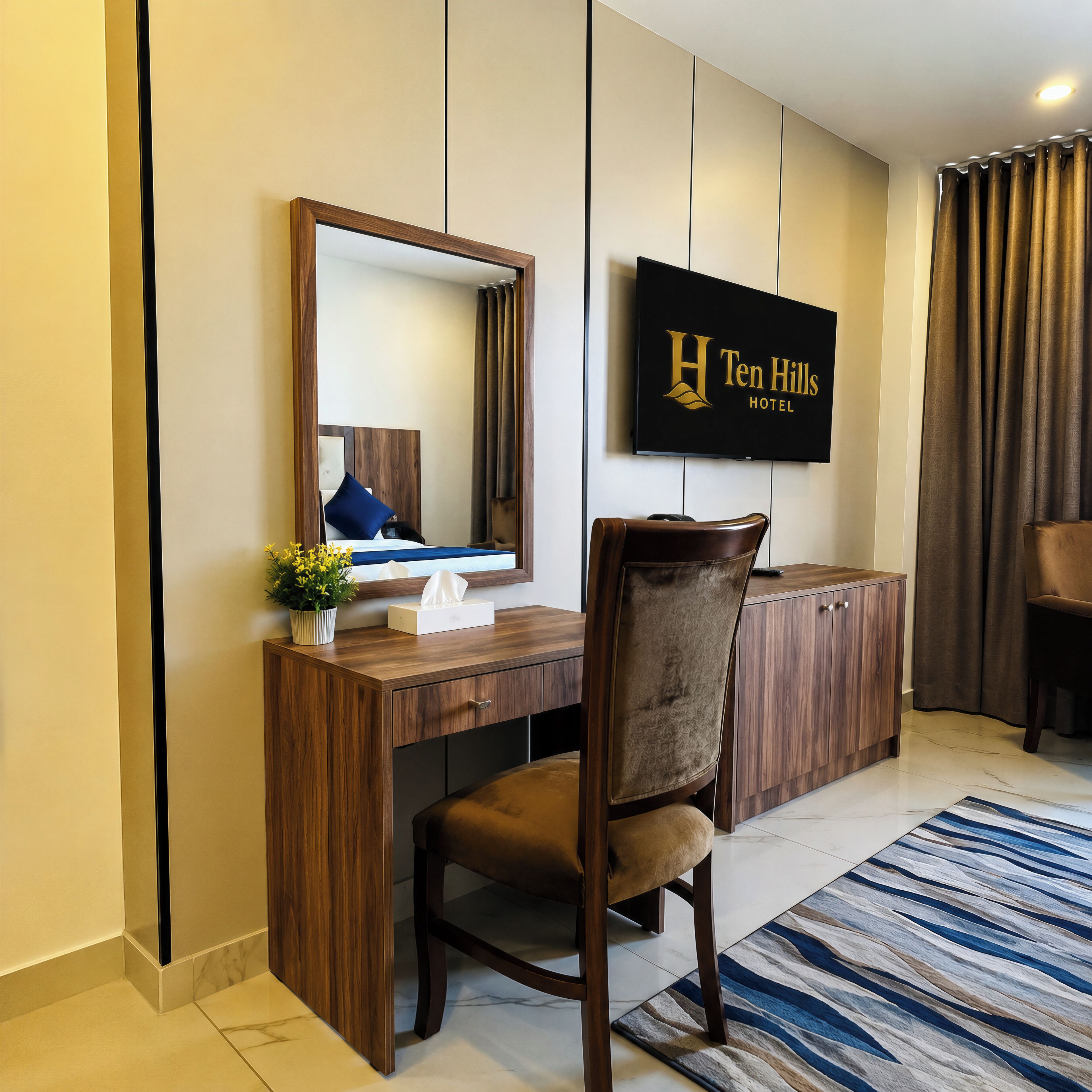 A hotel room with a wooden desk, a chair, a large mirror, a flat-screen TV, and a sign for 'Ten Hills Hotel'.