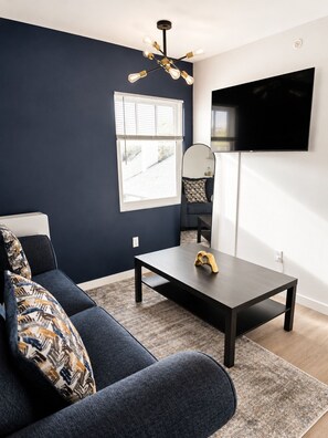 Un salon moderne avec un mur d’accent bleu foncé, un canapé agrémenté de coussins à motifs, une table basse, un téléviseur fixé au mur et une fenêtre avec des stores.