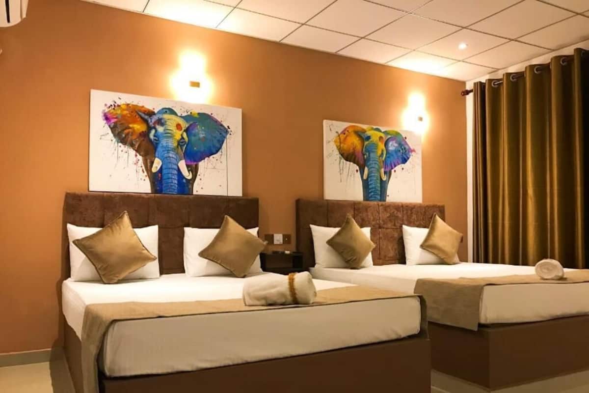 A hotel room with two beds, each with a headboard, white bedding, and beige pillows. There are two wall-mounted paintings of elephants.