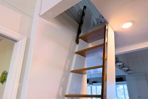 Eine Holzleiter lehnt an einer weißen Wand, eine Deckenleuchte und ein Fenster mit Blick.