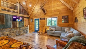 Ein Interieur einer Blockhütte mit Holzbalkendecke, einem aufgehängten Hirschgeweih, einer Steinwand, einem grauen Sofa und einem Holzesstisch.