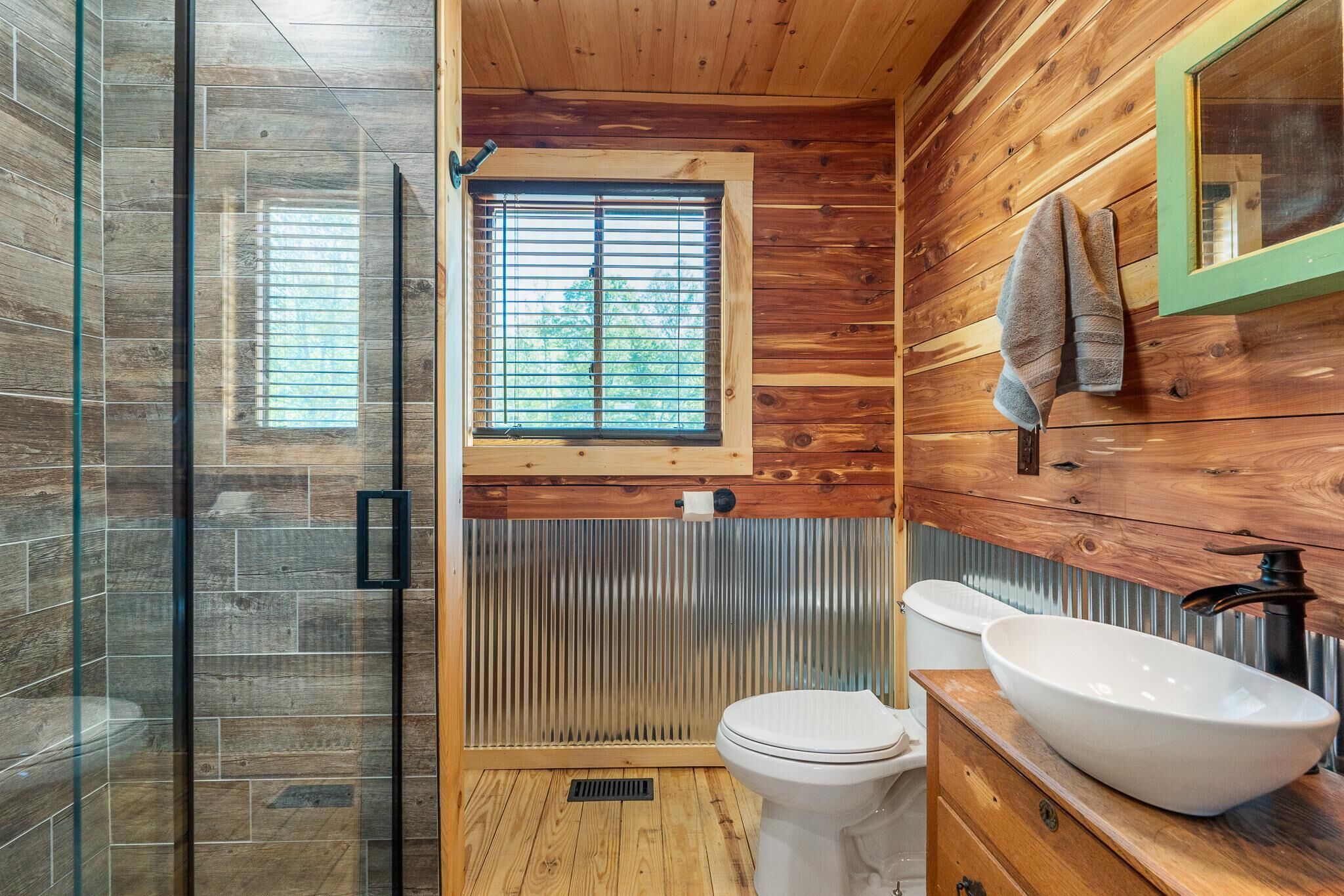 A bathroom with a wooden floor, a glass shower enclosure, a toilet, a sink with a basin, and a window with blinds.