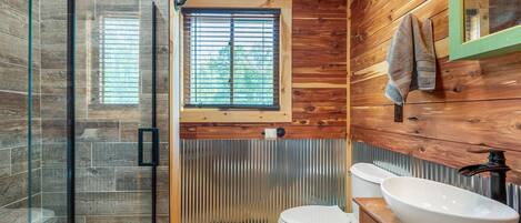 A bathroom with a wooden floor, a glass shower enclosure, a toilet, a sink with a basin, and a window with blinds.