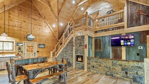 Ein rustikales Blockhaus-Interieur mit einem Holzesstisch, einem Steinkamin, einer Treppe und einem montierten Fernseher, der Streaming-Dienste anzeigt.