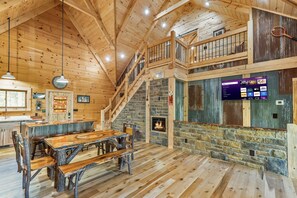 Interior rústico de una cabaña con una mesa de comedor de madera, una chimenea de piedra, una escalera y un televisor montado que muestra servicios de transmisión.