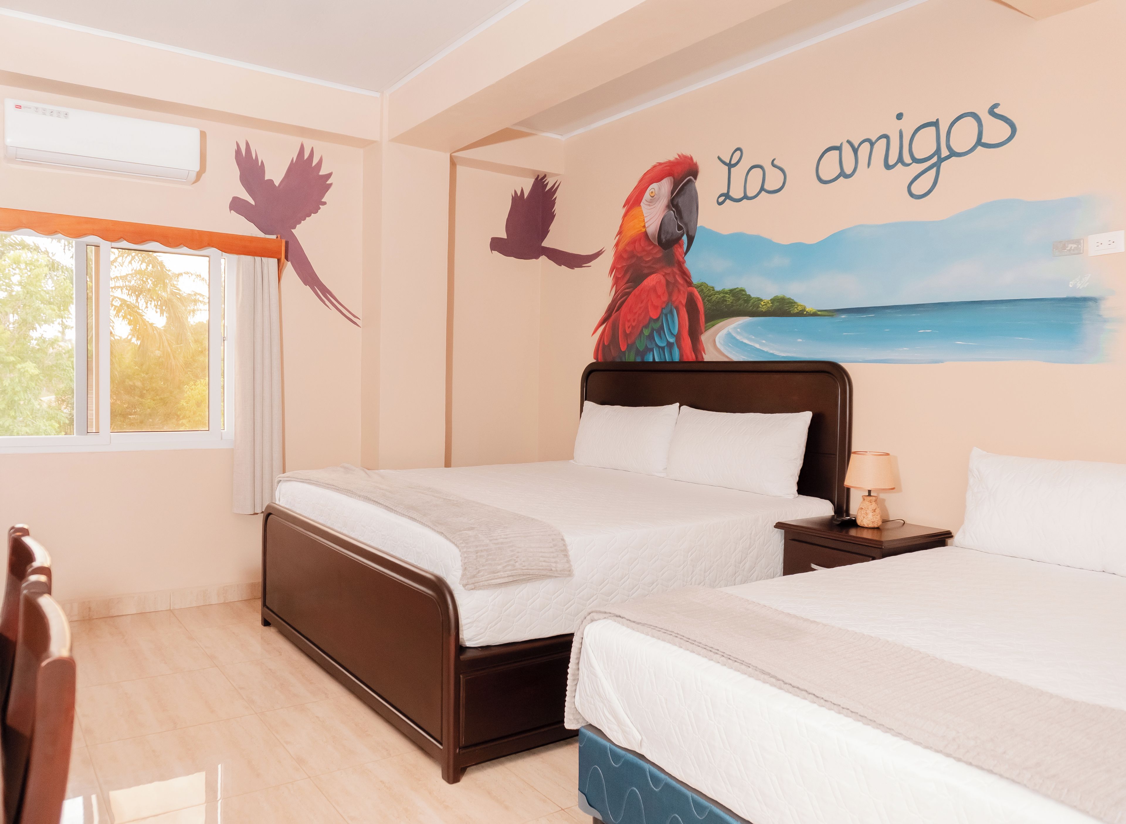 A hotel room with two beds, a mural of a parrot and a beach, and a window with curtains.