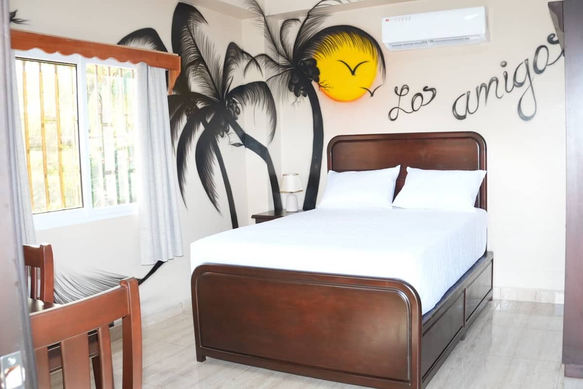 A bedroom with a bed, a chair, a wall with palm tree artwork, and a wall with the text 'Los amigos'.