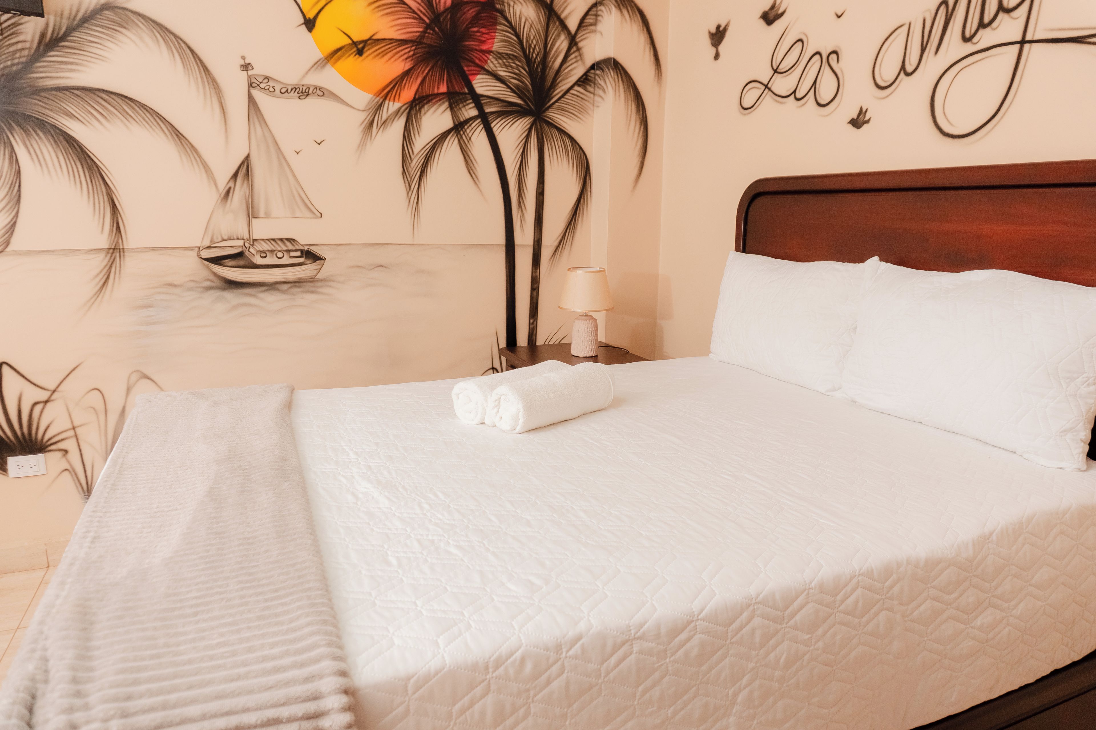 A neatly made bed with a white comforter and a rolled white towel, a wooden headboard, and a wall mural with a sailboat and palm trees.