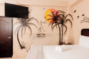 Habitación de hotel con cama, televisor montado en la pared y un mural de palmeras y un velero.