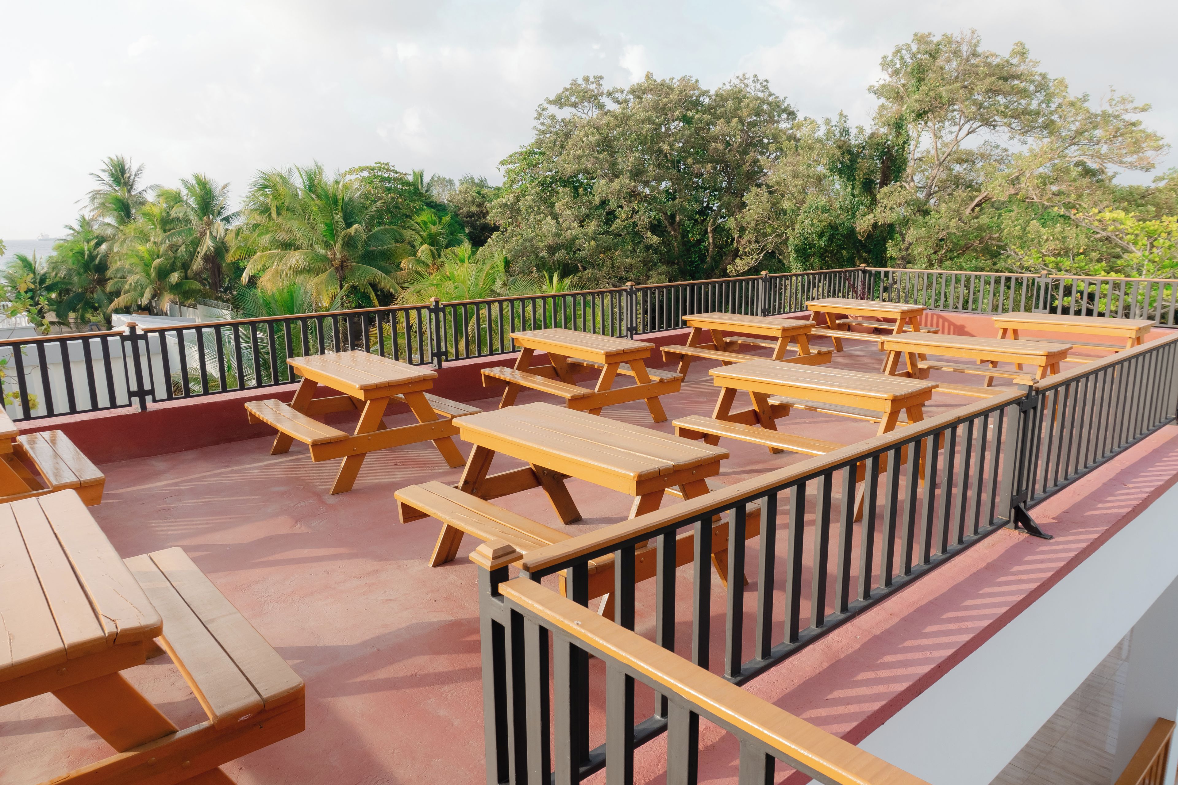 Outdoor wooden picnic tables arranged on a red deck with a black railing, overlooking a tropical landscape with palm trees and a body of water.