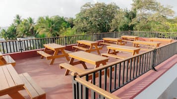 Mesas de picnic de madera al aire libre dispuestas en un piso rojo con barandilla negra, con vistas a un paisaje tropical con palmeras y una masa de agua.