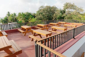Outdoor wooden picnic tables arranged on a red deck with a black railing, overlooking a tropical landscape with palm trees and a body of water.