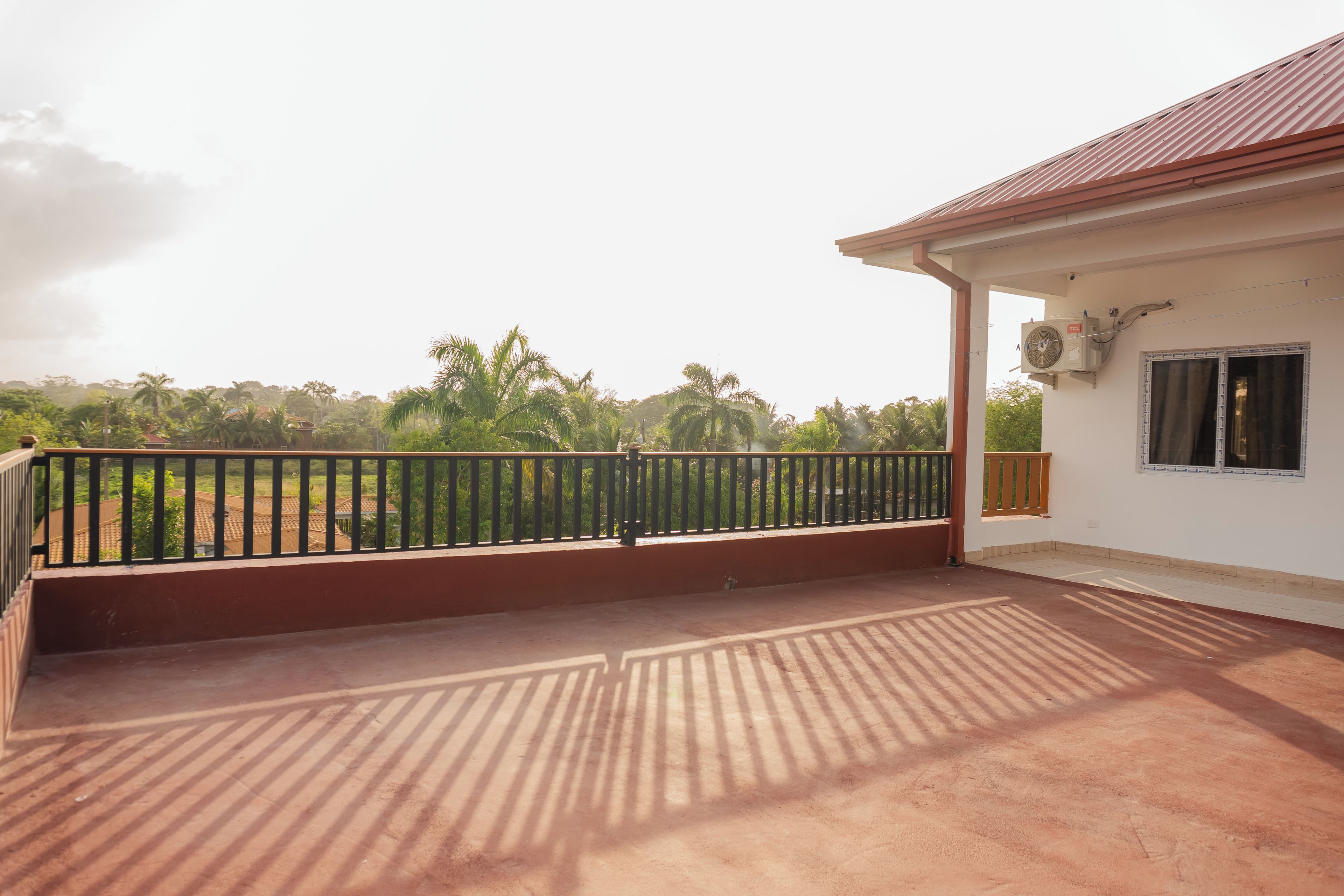 A balcony with a metal railing, a concrete floor, and a view of a green landscape with trees and a building in the distance.