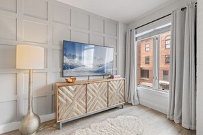 A modern living room with a large flat-screen TV, a decorative console, a floor lamp, and a view of a brick building through a window. - Location! Luxury Main St Condo - Walk to Town Lift (Park City)