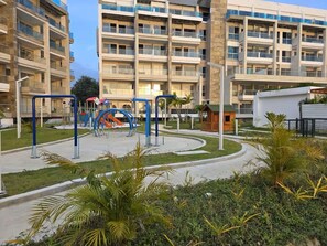 Ein Wohngebäude mit mehreren Balkonen, ein Spielplatz mit blau-orangen Geräten und eine angelegte Grünfläche mit Palmen.