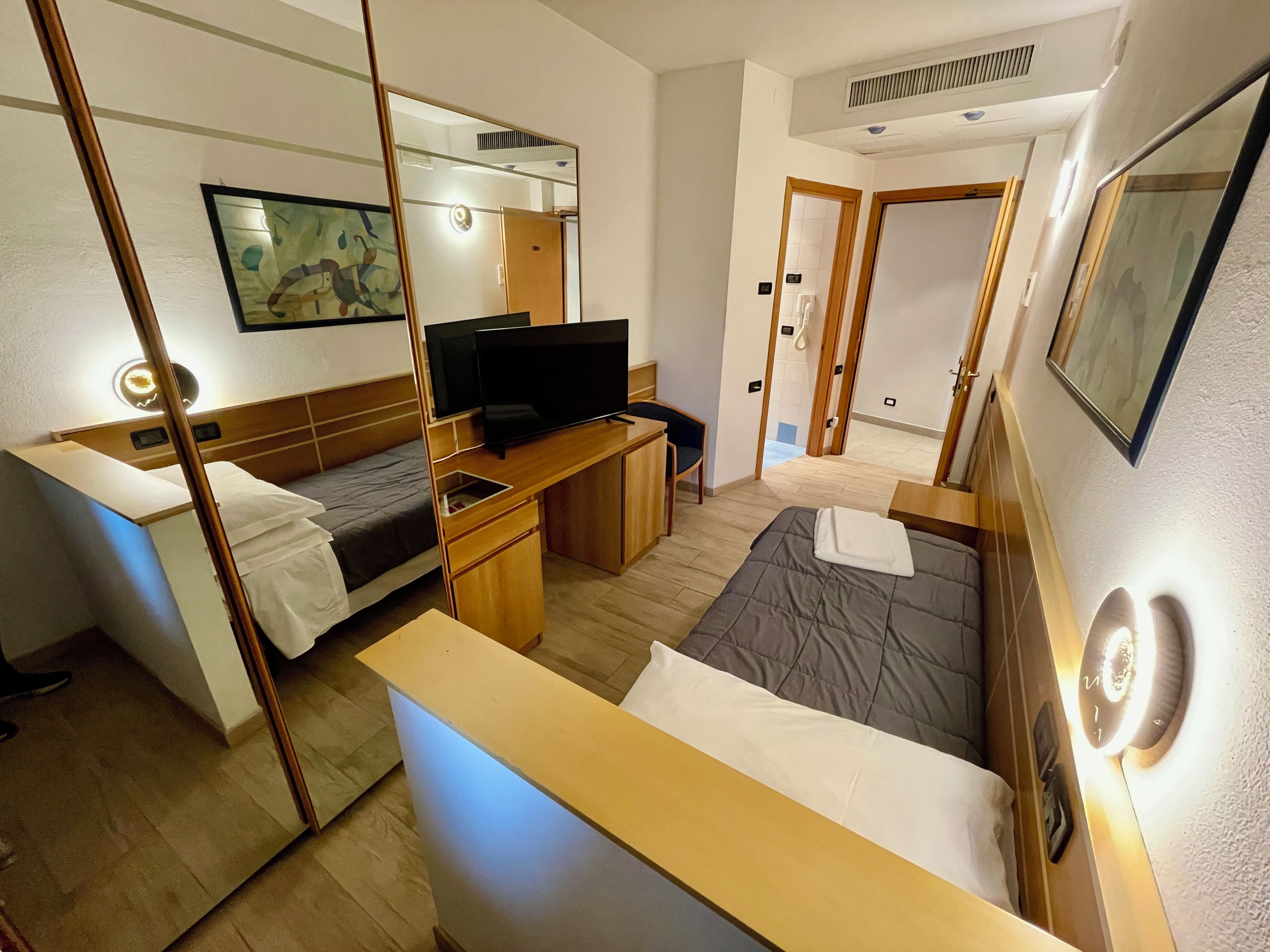 A hotel room with a bed, a desk with a television, a mirror, and a door leading to a bathroom.