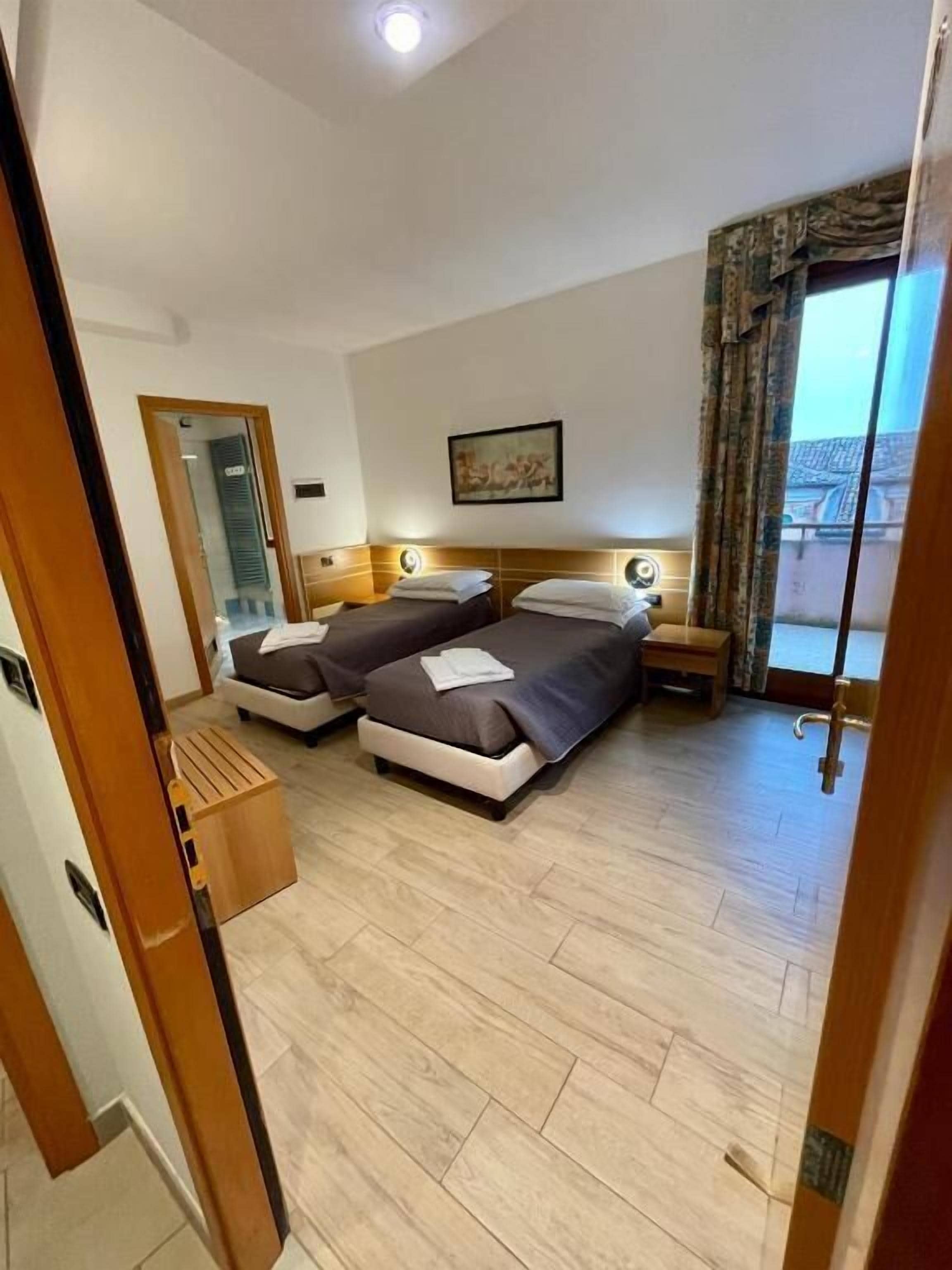 Ein Hotelzimmer mit zwei Betten, Holzboden, einem Fenster mit Vorhängen und einem Bild an der Wand.